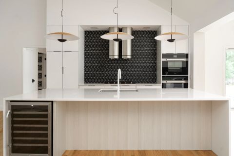Aiken kitchen with white quartz island, light wood paneling, wine cooler, black geometric tile backsplash, dual range hoods, and double ovens by Tekton Builds.