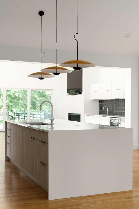 Aiken kitchen with L-shaped island, white quartz countertop, light wood cabinetry, mustard-yellow pendant lights, and sliding glass doors by Tekton Builds.