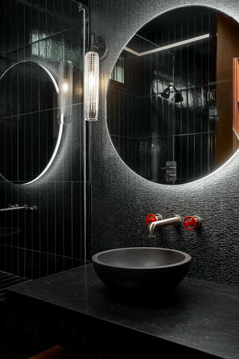Aiken bathroom with circular backlit mirror, dark concrete vessel sink, brushed metal faucet with red handles, and textured black tile wall by Tekton Builds.