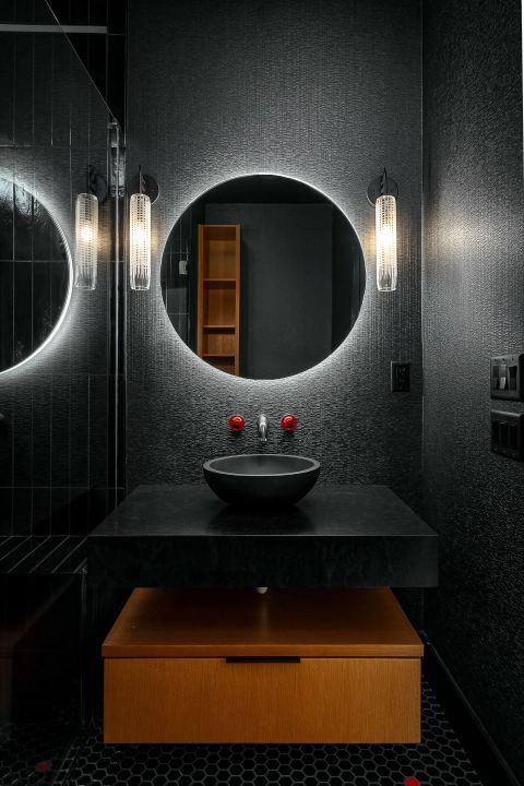 Aiken bathroom with dark textured walls, circular backlit mirror, floating black vanity with vessel sink, and hexagonal mosaic floor tiles by Tekton Builds.