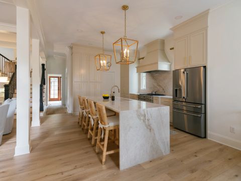 Greenville kitchen with marble island, brass faucet, cane bar stools, cream shaker cabinets with gold hardware, and veined marble backsplash by Tekton Builds.