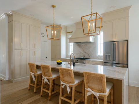 Greenville kitchen with elongated marble island, gold faucet, cane bar stools, geometric brass pendant lights, cream shaker cabinets, and stainless steel range by Tekton Builds.
