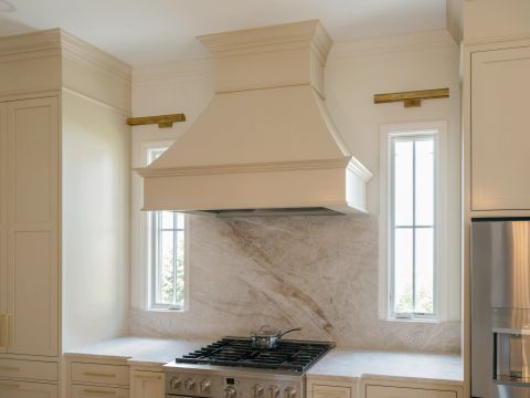 Greenville kitchen with custom cream range hood, stainless steel range, marble backsplash with brown and grey veining, and brass picture lights by Tekton Builds.