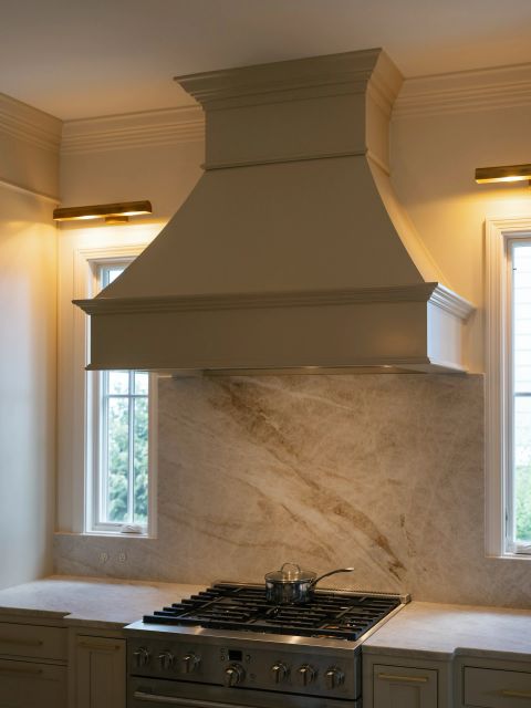 Greenville kitchen with stainless steel range, custom beige range hood, marble backsplash with brown-grey veining, and brass lighting by Tekton Builds.
