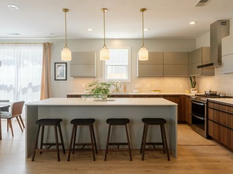 Greenville kitchen with four walnut bar stools, light gray island, taupe upper cabinets, stainless steel range with chimney hood, and gold pendant lighting by Tekton Builds.