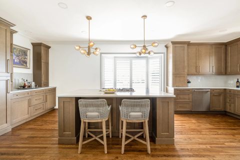 Greenville kitchen with warm brown cabinetry, central island with seating for two, sputnik-style chandeliers with amber glass globes, white quartz countertops, and plantation shutters by Tekton Builds.