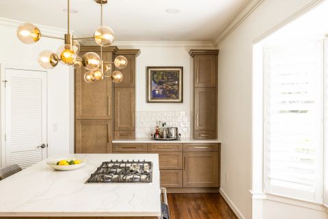 Greenville kitchen with white quartz island with integrated gas cooktop, warm brown cabinetry with gold bar pulls, subway tile backsplash, brass chandelier with amber glass, and plantation shutters by Tekton Builds.