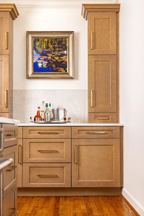 Greenville wet bar with light brown wood cabinetry, gold hardware, white countertop, subway tile backsplash, and water lily painting by Tekton Builds.