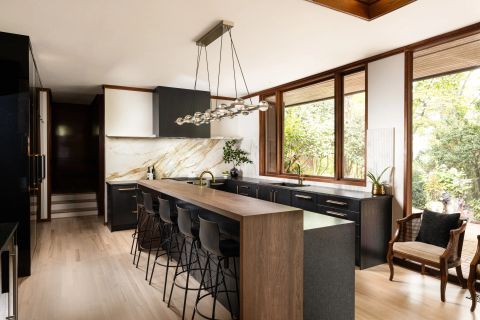 Greenville kitchen with dark cabinetry, wooden island with seating for six, marble backsplash, brushed gold gooseneck faucets, and linear chandelier by Tekton Builds.