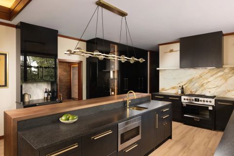 Greenville kitchen with black cabinetry, long brass hardware, multi-level island with dark stone countertop, marble backsplash, and linear brass chandelier by Tekton Builds.