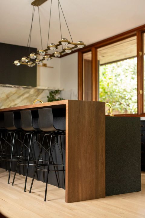 Greenville kitchen island with warm wood top and side panels, black bar stools, linear brass chandelier, and wood-framed windows by Tekton Builds.
