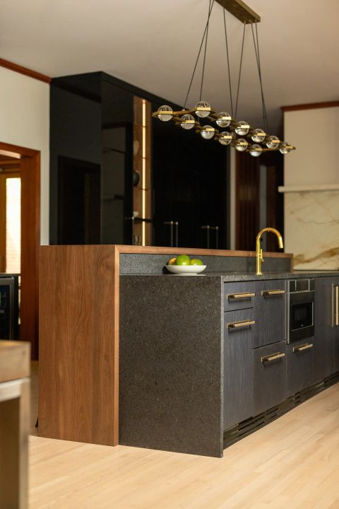 Greenville kitchen island with dark cabinetry, reddish-brown wood accent panel, golden gooseneck faucet, integrated microwave drawer, and linear chandelier by Tekton Builds.