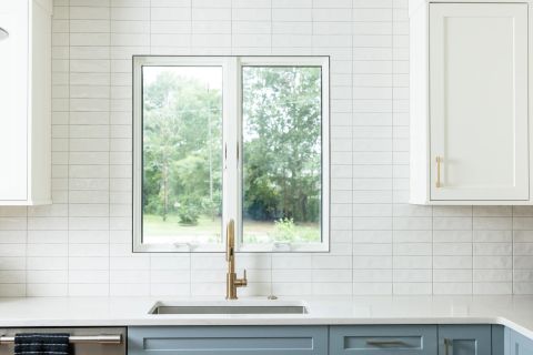 Taylors kitchen sink area with double window, stainless steel undermount sink, brushed gold gooseneck faucet, light blue-grey shaker cabinets, and subway tile backsplash by Tekton Builds.