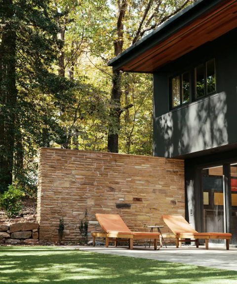 Two-story home addition with dark siding, wood soffit, stacked-stone privacy wall, and patio lounge chairs in a wooded backyard.