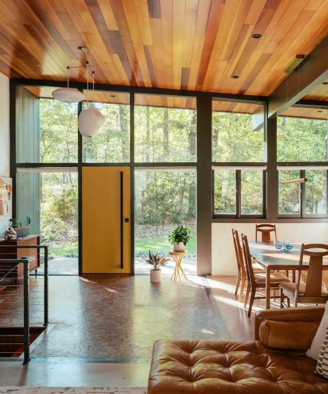Renovated living and dining space with wood-plank ceiling, floor-to-ceiling windows, yellow entry door, and polished concrete floors.